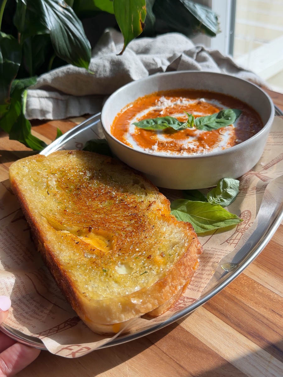 Bowl of roasted tomato soup with basil, served with a pesto grilled cheese on a tray in natural light.
