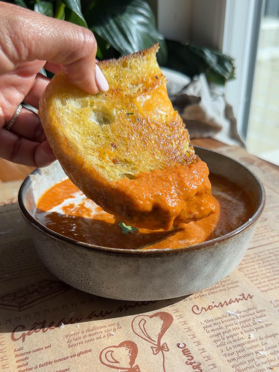 Dipping toasted pesto-grilled cheese into creamy tomato soup in a rustic ceramic bowl on a wooden table.