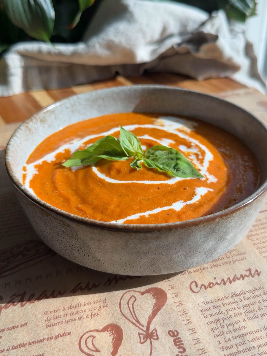 Roasted tomato soup topped with fresh basil and cream swirl in a ceramic bowl on wooden table, sunlight and shadows.