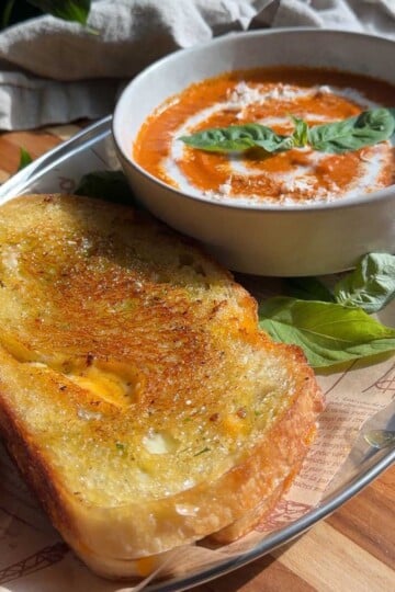 Bowl of roasted tomato soup with basil, served with a pesto grilled cheese on a tray in natural light.