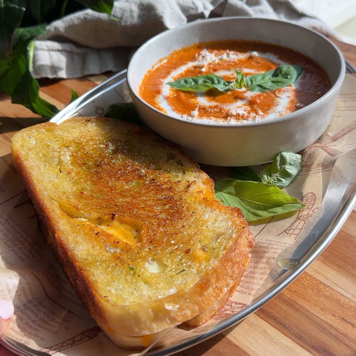 Bowl of roasted tomato soup with basil, served with a pesto grilled cheese on a tray in natural light.