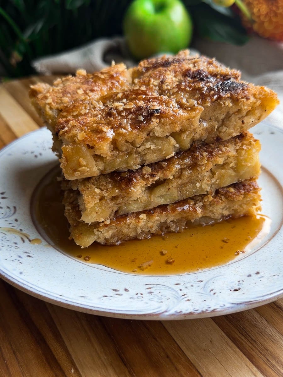 Sheet pan apple crumble pancakes stacked on a plate with syrup drizzle, wooden background.