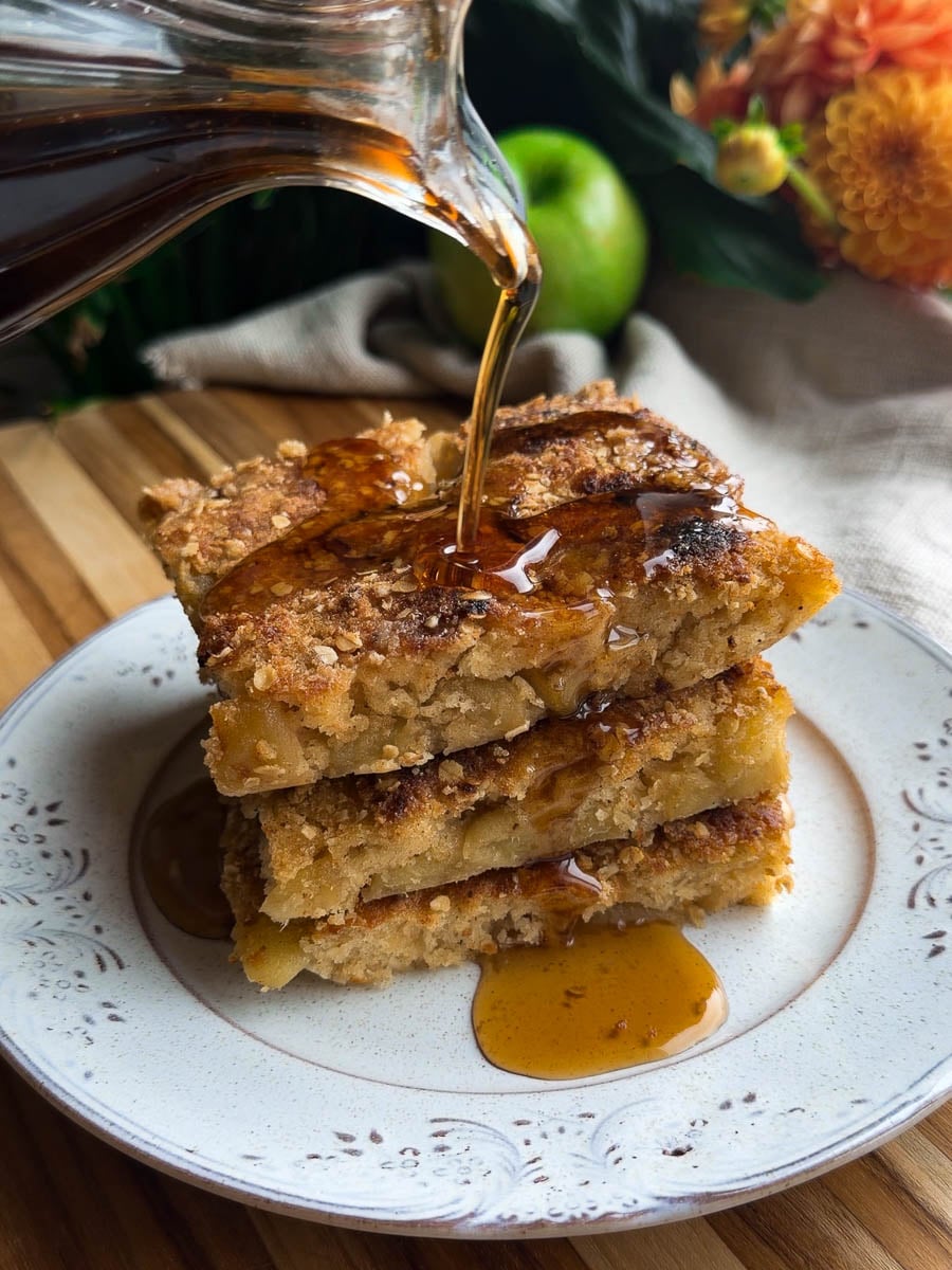 Stacked sheet pan apple crumble pancakes, with syrup pour on a decorative plate, green apple and flowers in background.