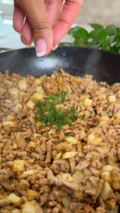 Sprinkling dill on cooked minced chicken and onions in a skillet.
