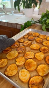 Baked potato slices on a tray held by a gloved hand in a kitchen setting.