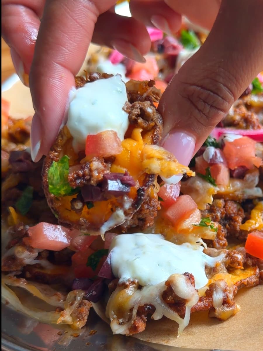 Hand holding loaded nachos topped with cheese, meat, sour cream, tomatoes, and herbs.