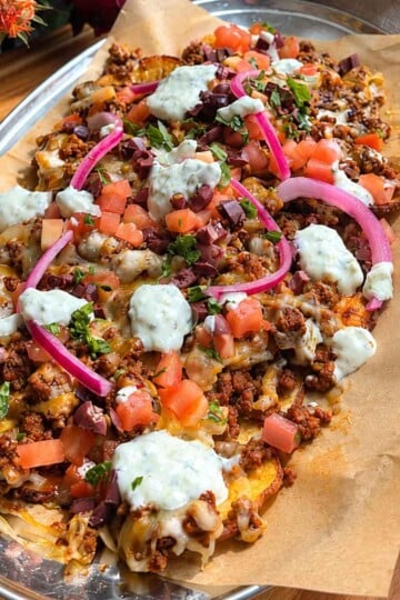 Greek loaded potato nachos with toppings including cheese, beef, tomatoes, olives, onions, and sour cream on a wooden table.