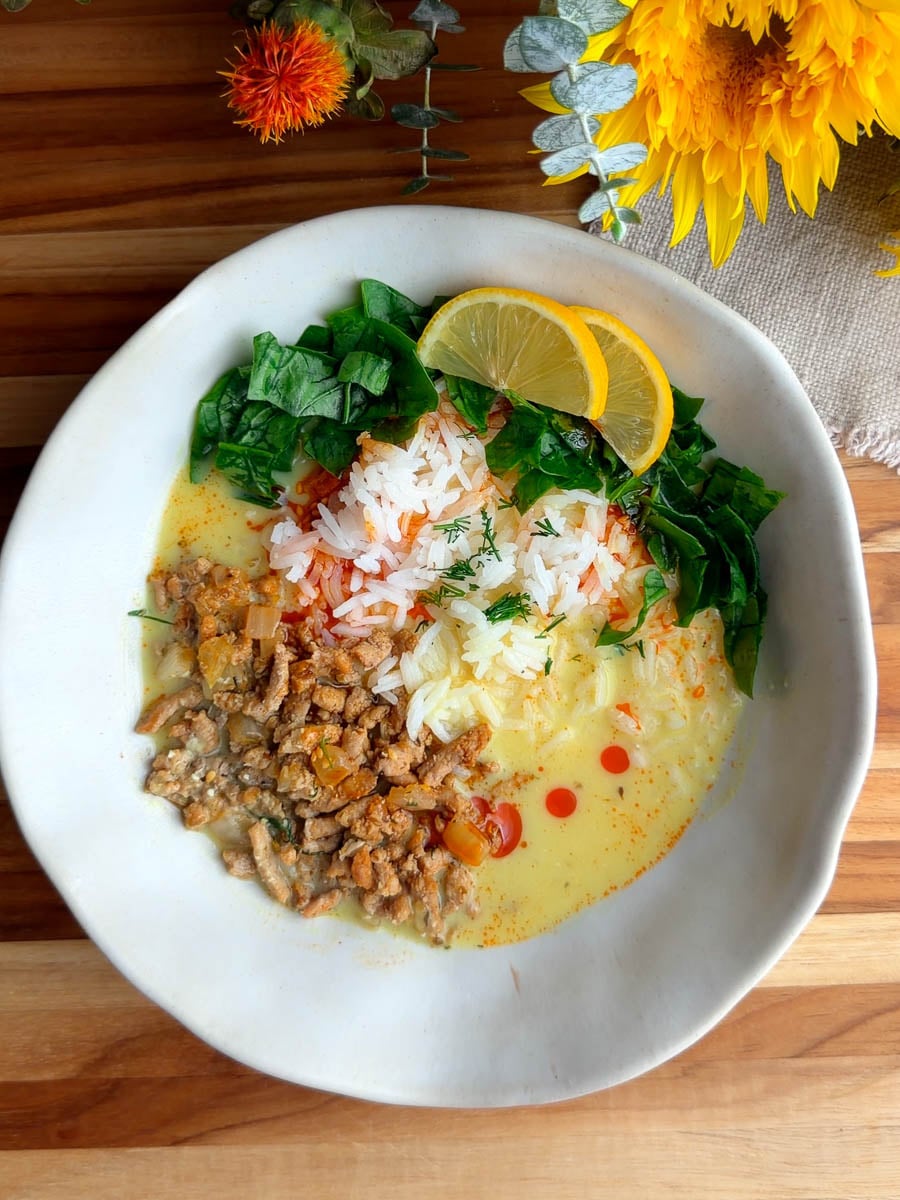 Bowl of Mediterranean brothy rice and chicken with spinach, minced meat, and lemon slices in a creamy sauce on a wooden table.