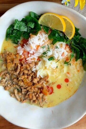 Bowl of Mediterranean brothy rice and chicken with spinach, minced meat, and lemon slices in a creamy sauce on a wooden table.