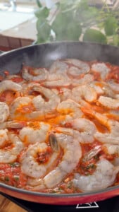 Shrimp cooking in a pan with tomato sauce and herbs, steam rising.