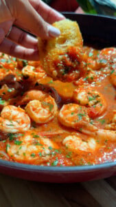 Shrimp in sauce with bread being dipped, garnished with herbs in a skillet.