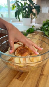 Hand mixing seasoned potato slices in a glass bowl on a wooden kitchen counter.