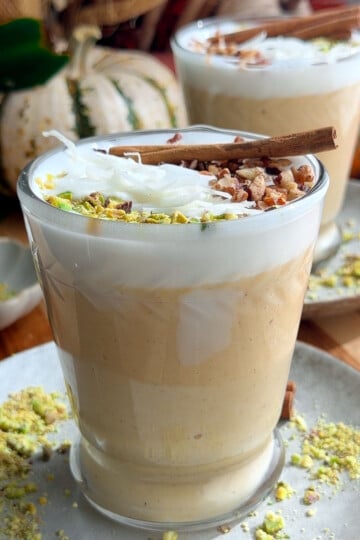 Creamy pumpkin sahlab topped with coconut, pistachios, chopped nuts, and cinnamon stick in a glass cup