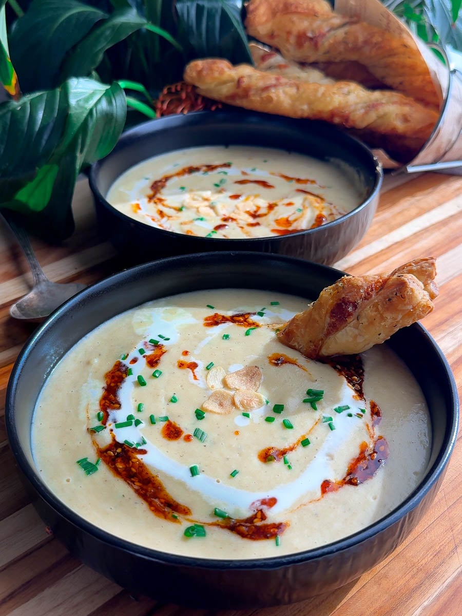Creamy roasted garlic soup topped with chives, chili oil, and garlic, served with puff pastry bread sticks in black bowls