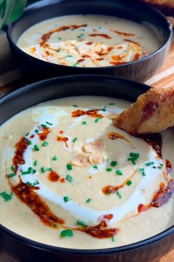 Creamy roasted garlic soup with chives, chili oil, and a crispy bread piece in a black bowl.