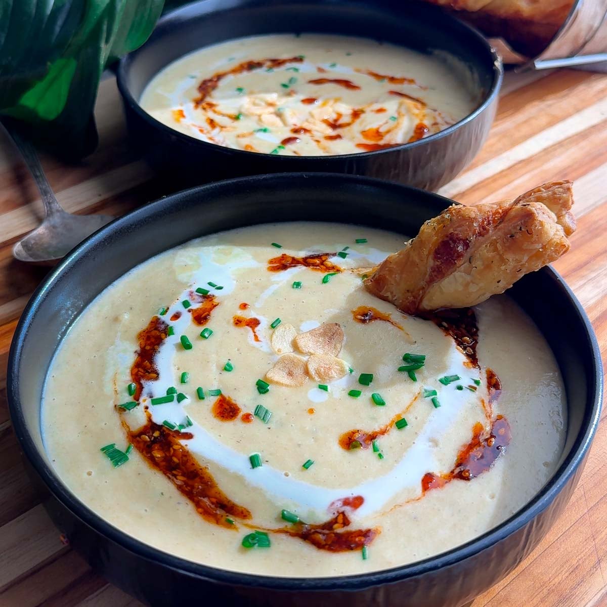 Creamy roasted garlic soup with chives, chili oil, and a crispy bread piece in a black bowl.